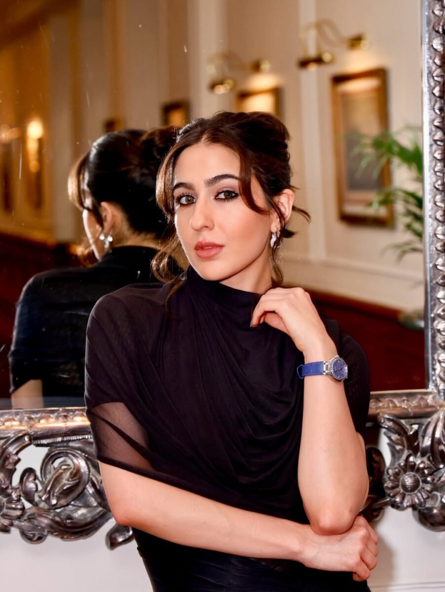 Sara Ali Khan posing in a chic, black draped high-neck dress with sheer sleeves, styled with a blue watch, stud earrings, and a sleek hairstyle, embodying elegant celebrity fashion.